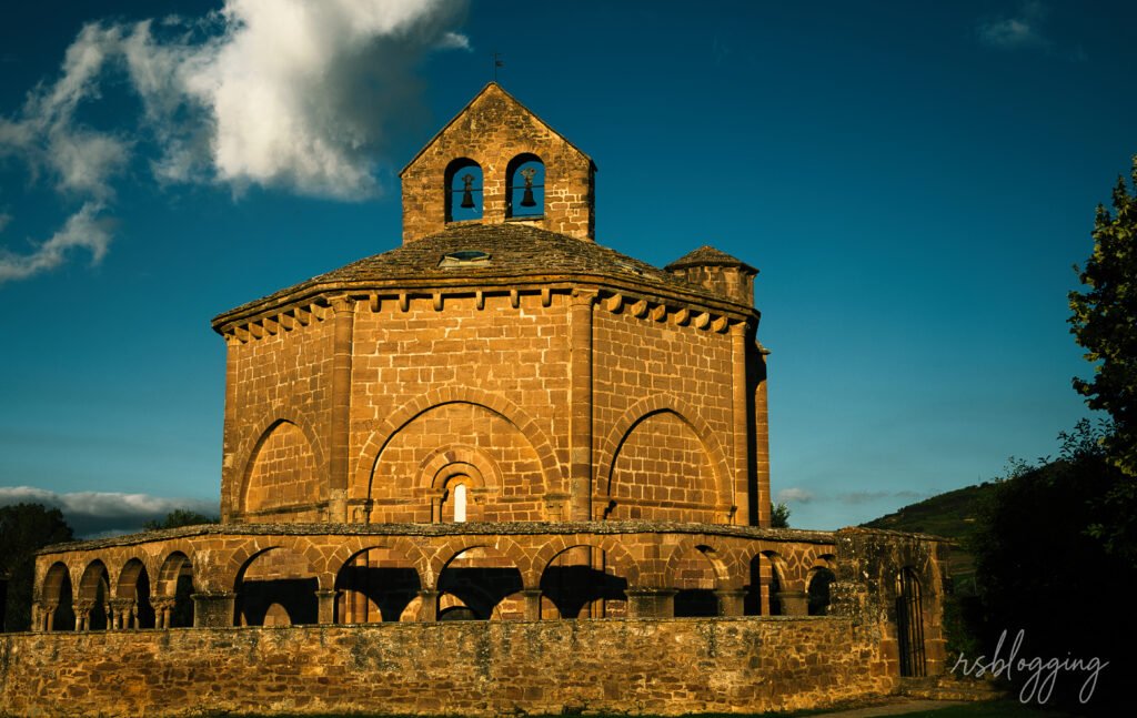 Ermita de Santa María de Eunate iluminada por el sol de la tarde, con cielo azul intenso y nubes al fondo